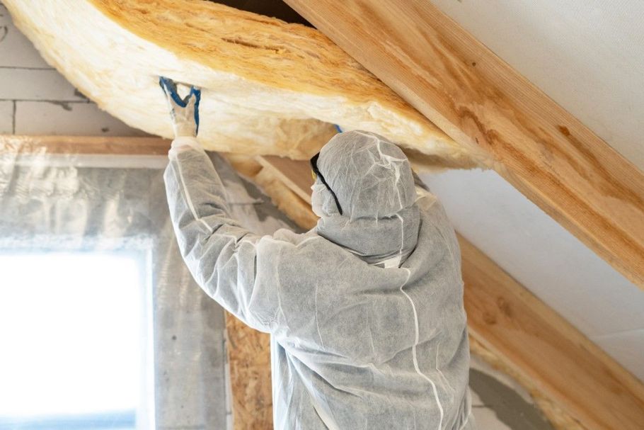 Person in a gray coverall insulation suit spray foaming attic rafters near a window