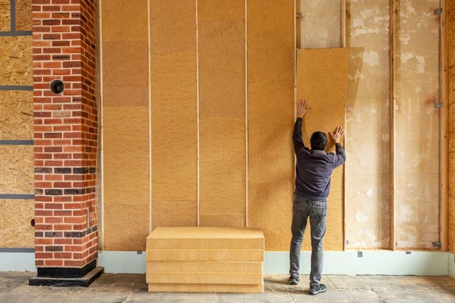 Person installing large tan wall panels beside a brick column in an unfinished room.