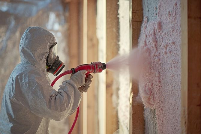 Worker in white protective suit spray-foaming insulation into a wall cavity during construction