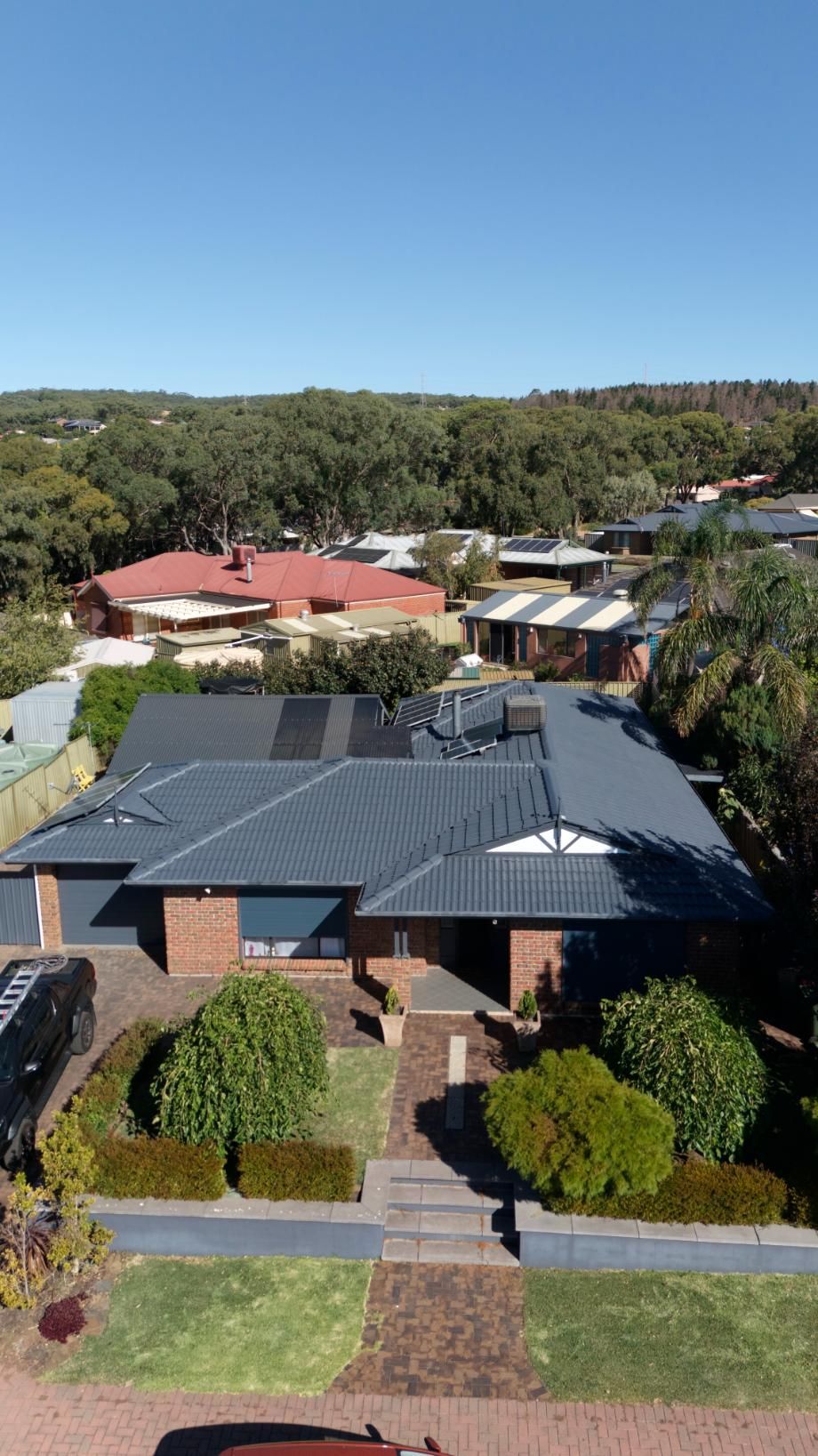 Suburban house with dark roof and solar panels, seen from above with trees and neighboring homes.