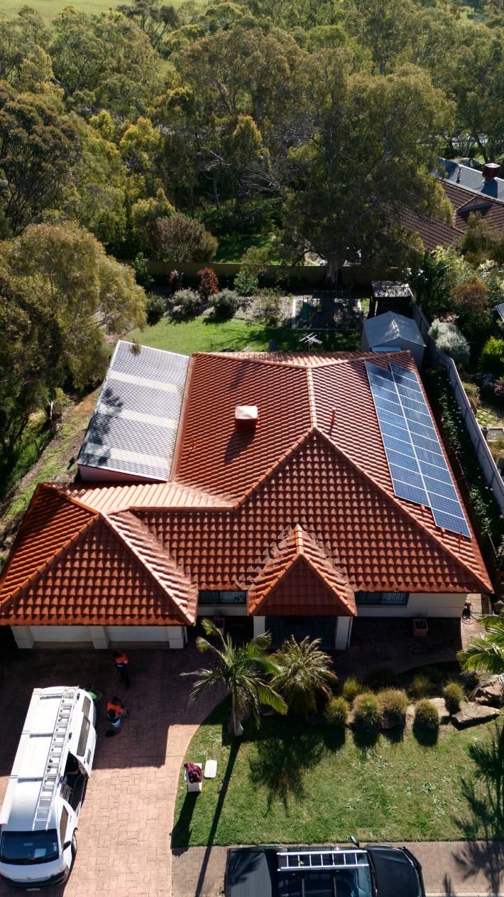 Aerial view of a house with a red-tile roof, solar panels, and a driveway surrounded by trees.