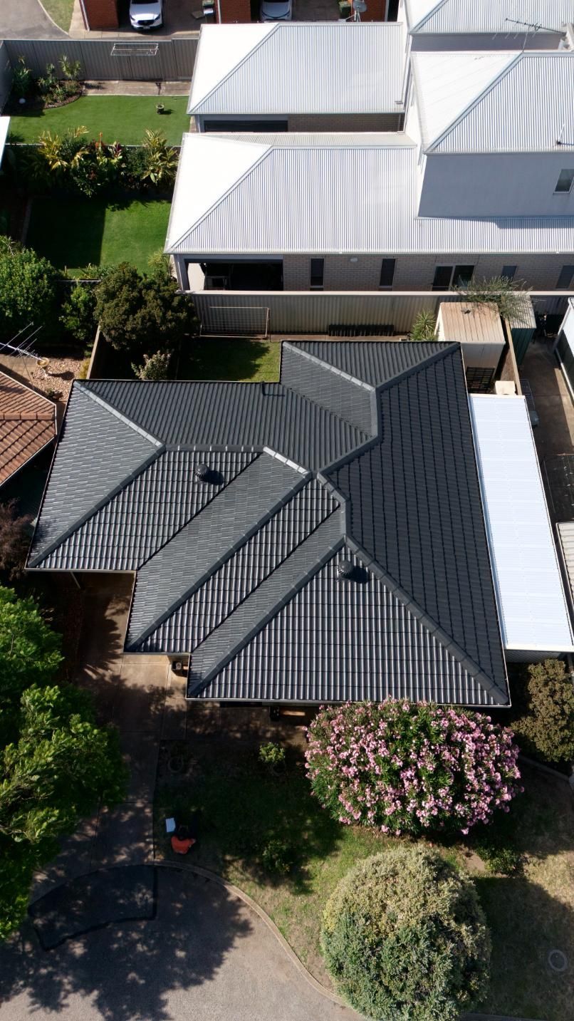 Aerial view of a dark gray roofed house with surrounding trees and neighboring white-roofed homes