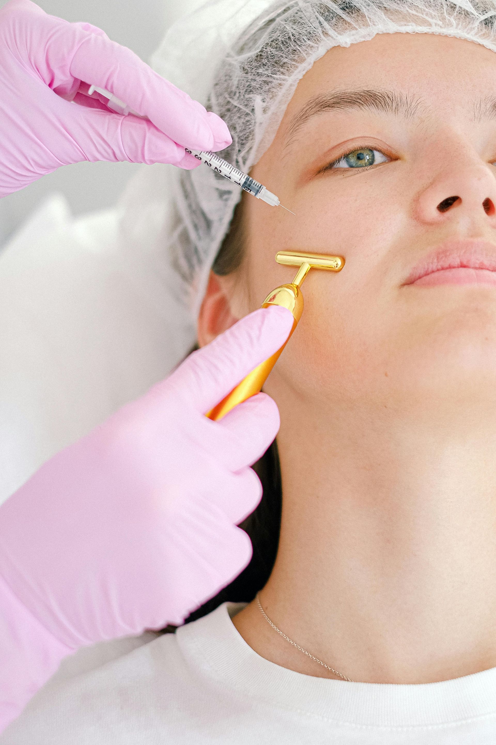 A person receiving a facial treatment. Pink-gloved hands hold a syringe and a golden facial massager near the face.