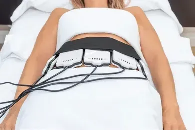 Woman lying down with a black belt device attached to her abdomen, connected to wires. She is on a white table, likely undergoing a medical procedure.