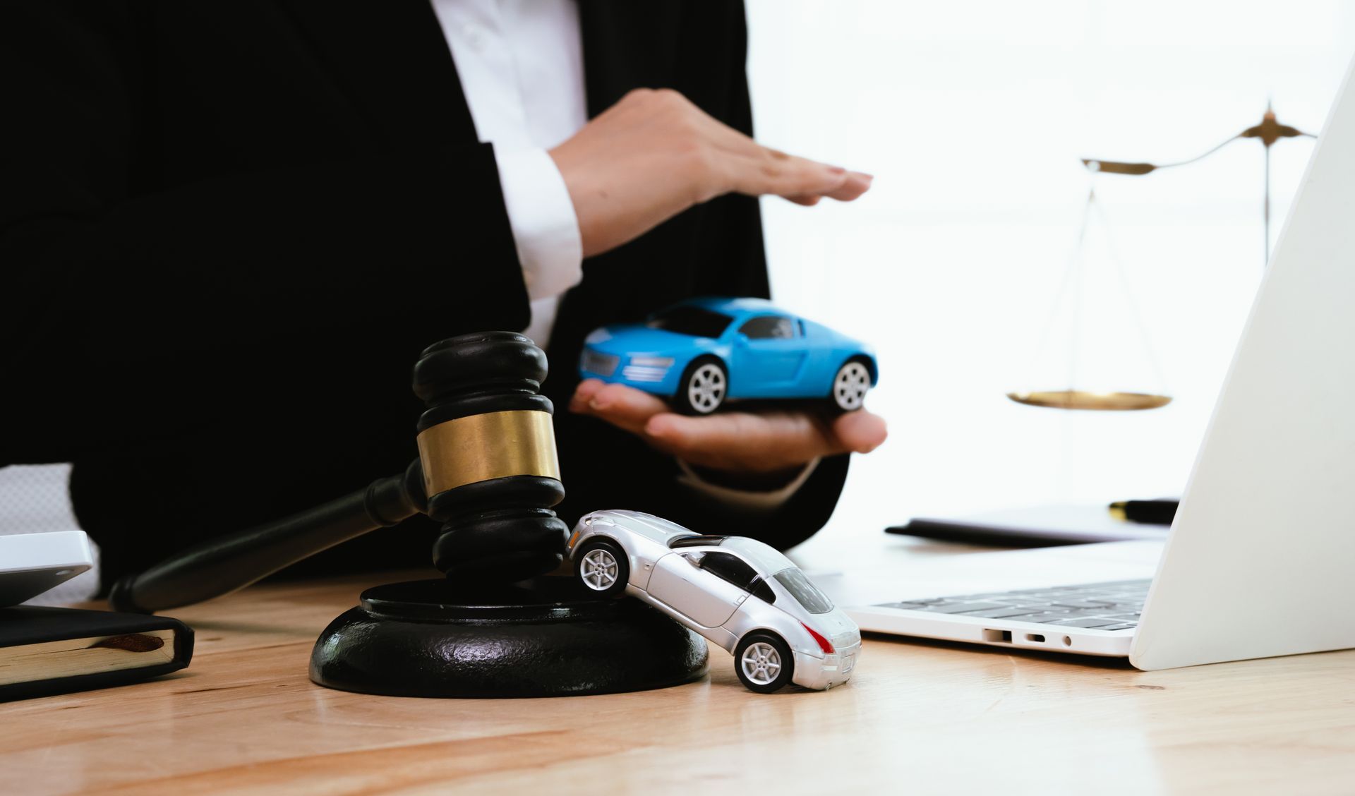 A lawyer in a suit holds a blue toy car beside a gavel and scale.