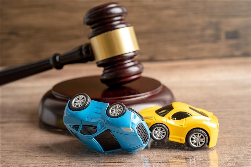 Two toy cars simulate a car accident, in front of a wooden gavel, on top of a wooden surface.