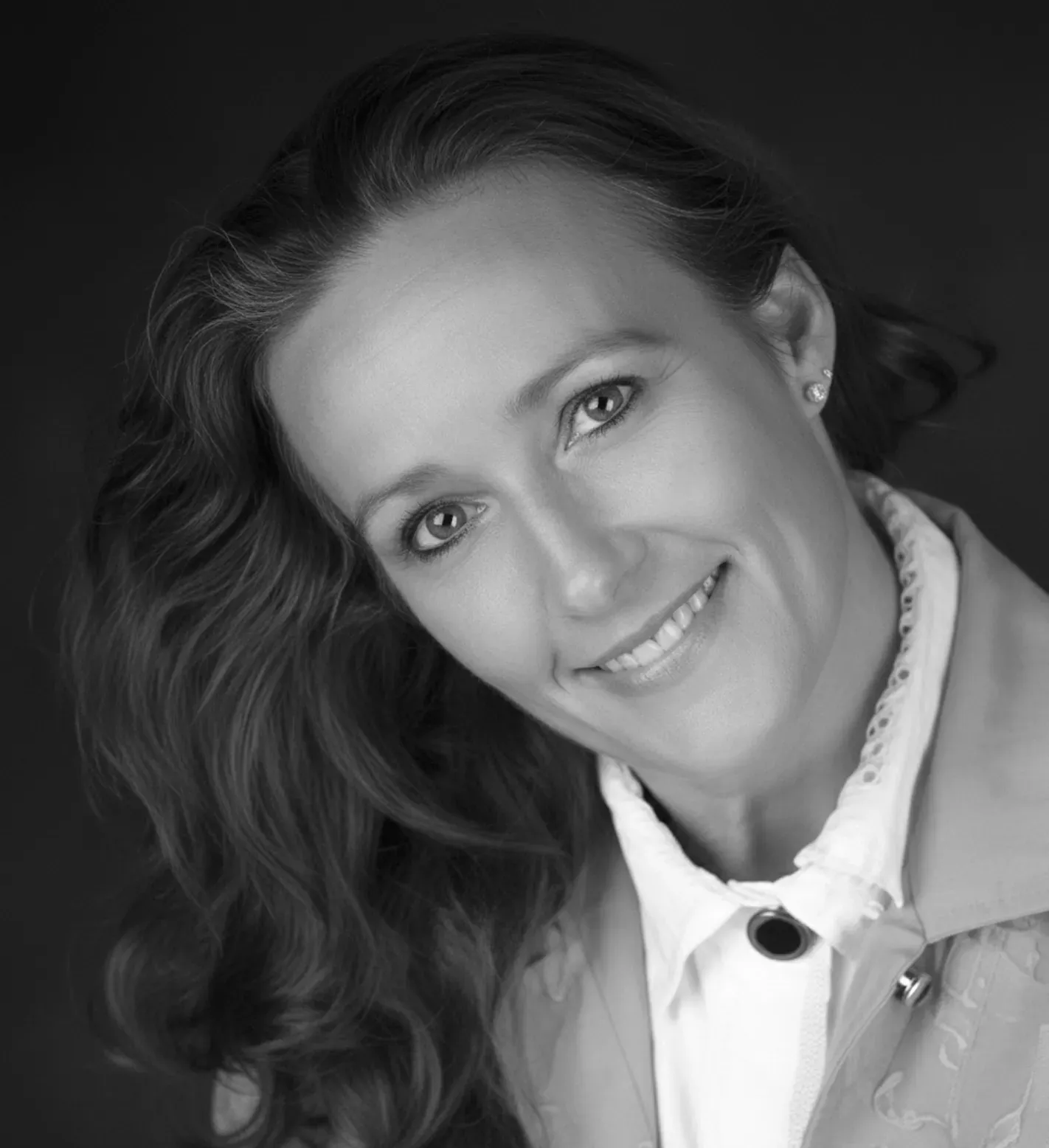 Woman with long wavy hair smiles, wearing a jacket and button-down shirt, head tilted, against a dark backdrop.