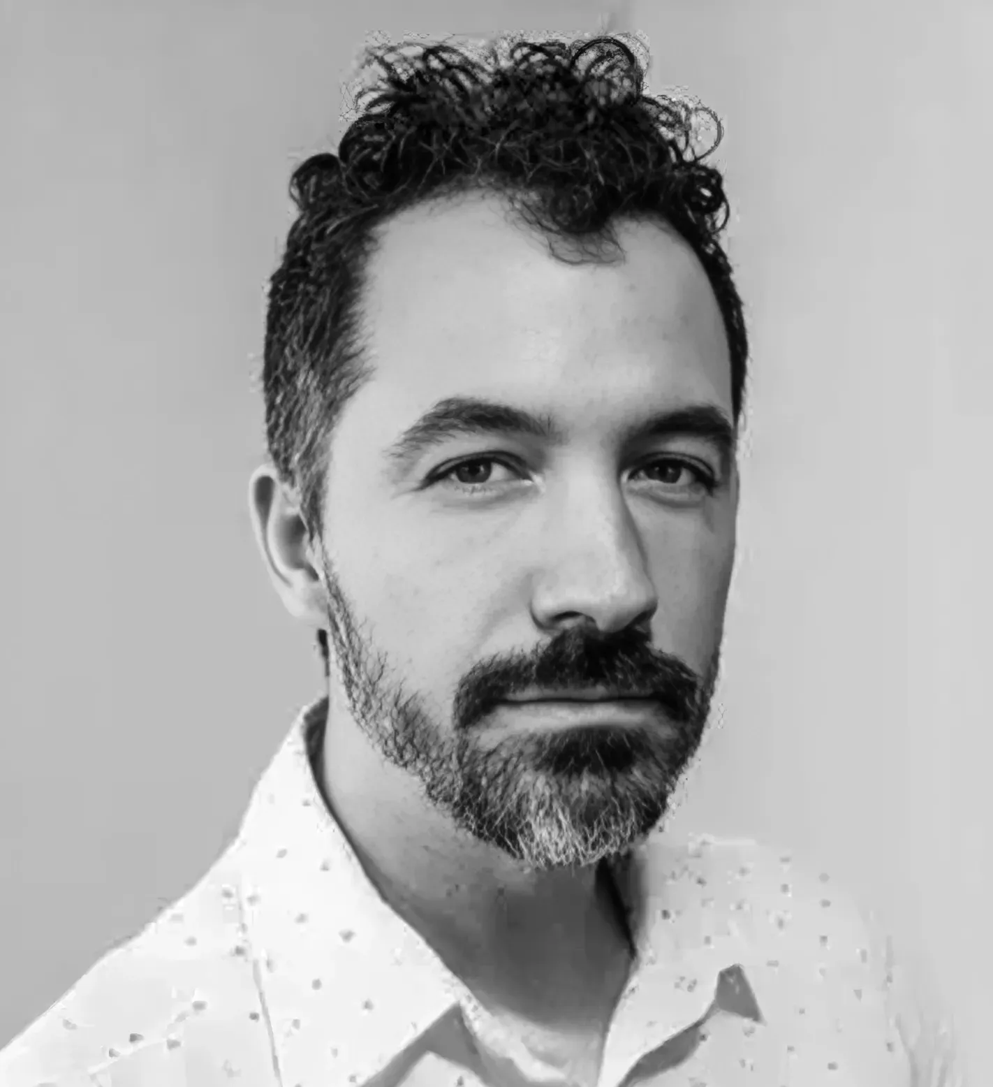 Man with dark hair and beard, wearing a patterned shirt, looking forward with a neutral expression.