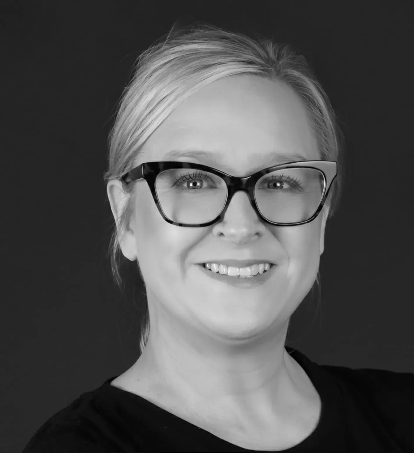 Woman with glasses smiling, black top, against a dark background.