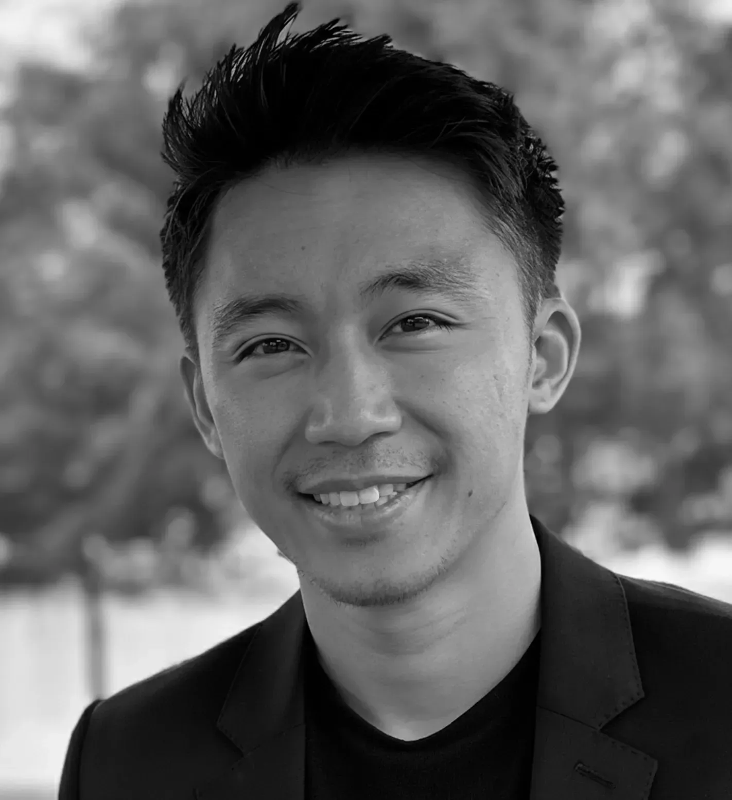 Smiling man in a dark blazer and black shirt, outdoors with blurred background.