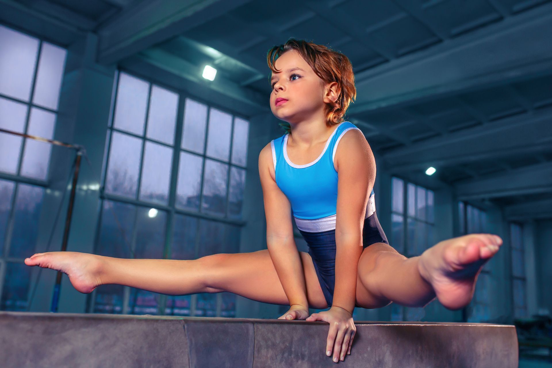 Gymnast in blue leotard performs split on a beam, hands down for support, in a gym.