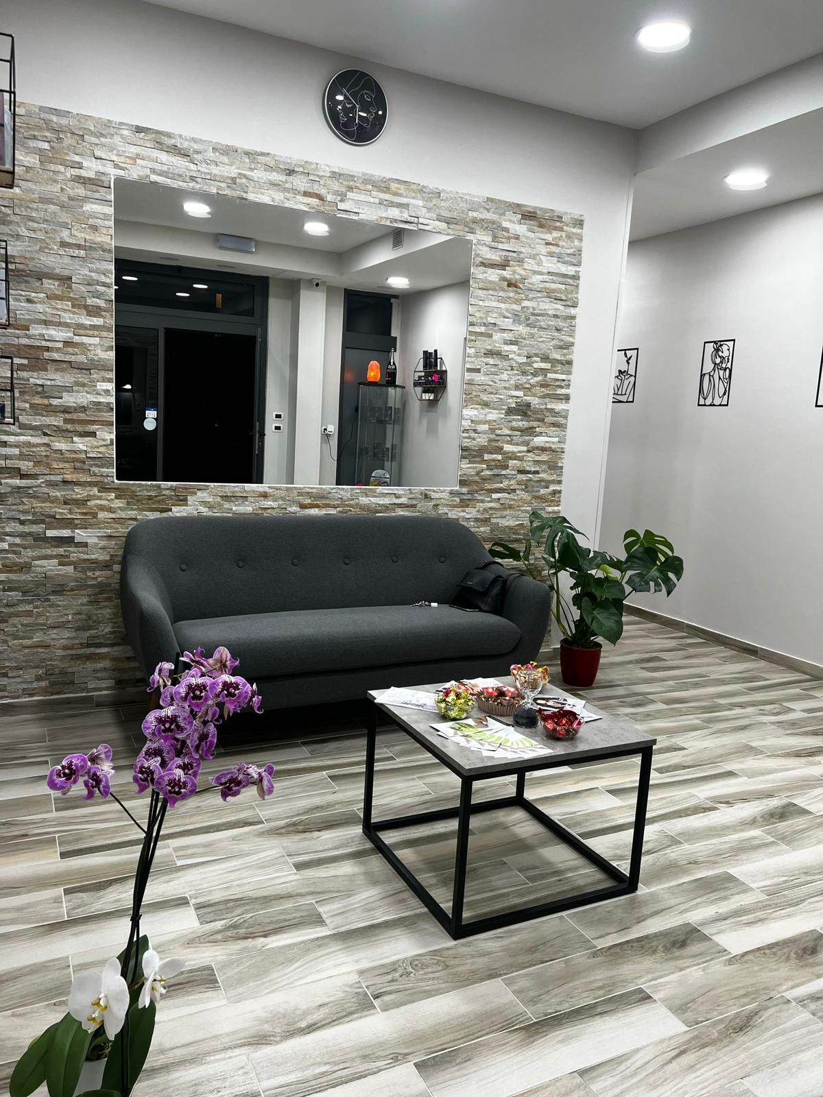 Accogliente sala d'attesa con divano grigio, tavolino, parete in pietra, specchio e piante in vaso.