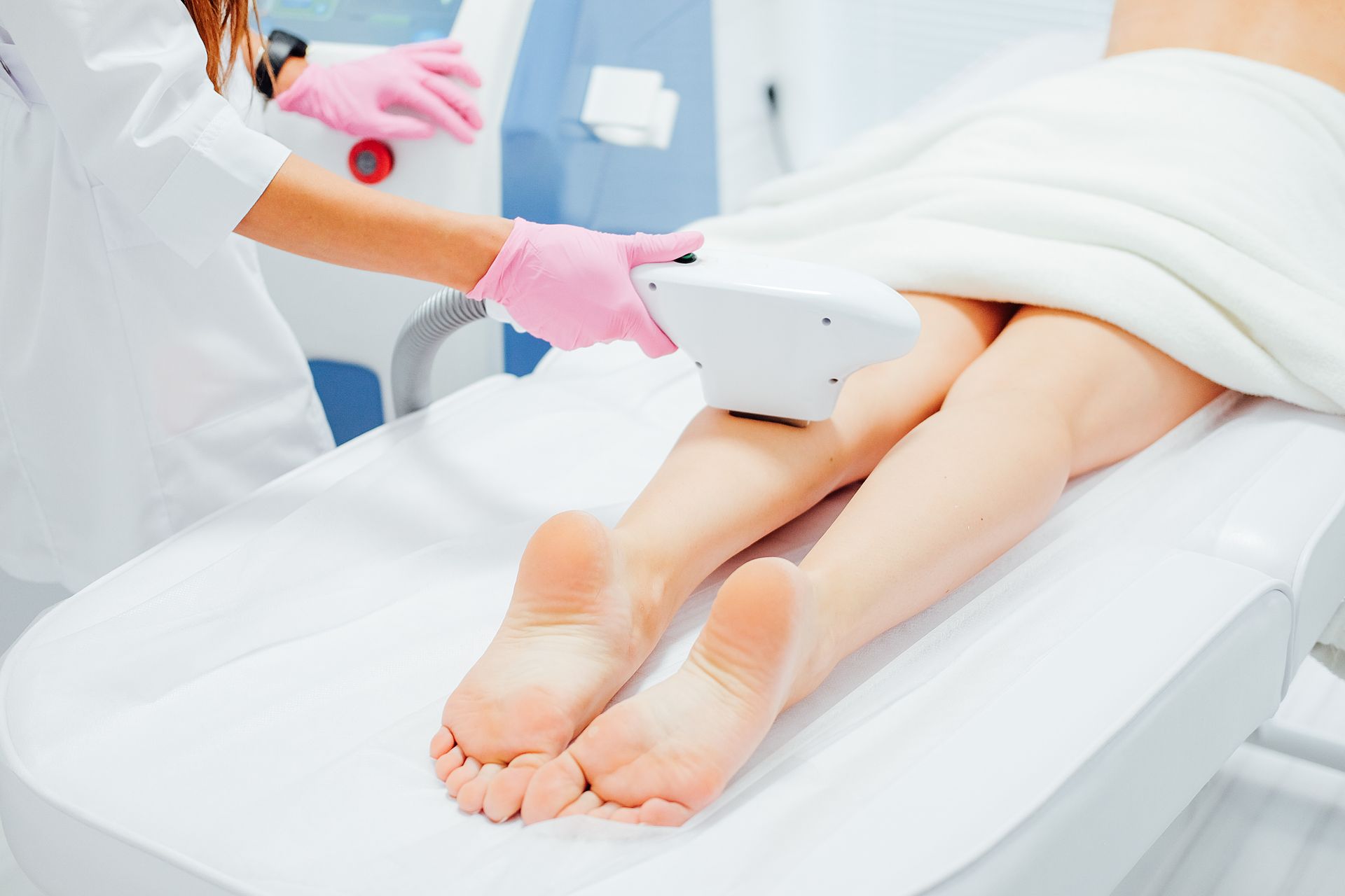Gambe su un lettino medico sottoposte a trattamento di epilazione laser. Una persona che indossa guanti rosa tiene in mano il dispositivo.