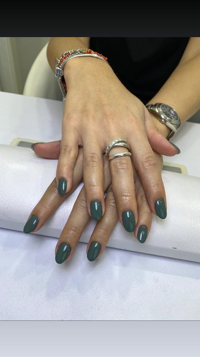 Mani con unghie ovali, verde scuro, appoggiate su una superficie bianca, con bracciali e anelli.