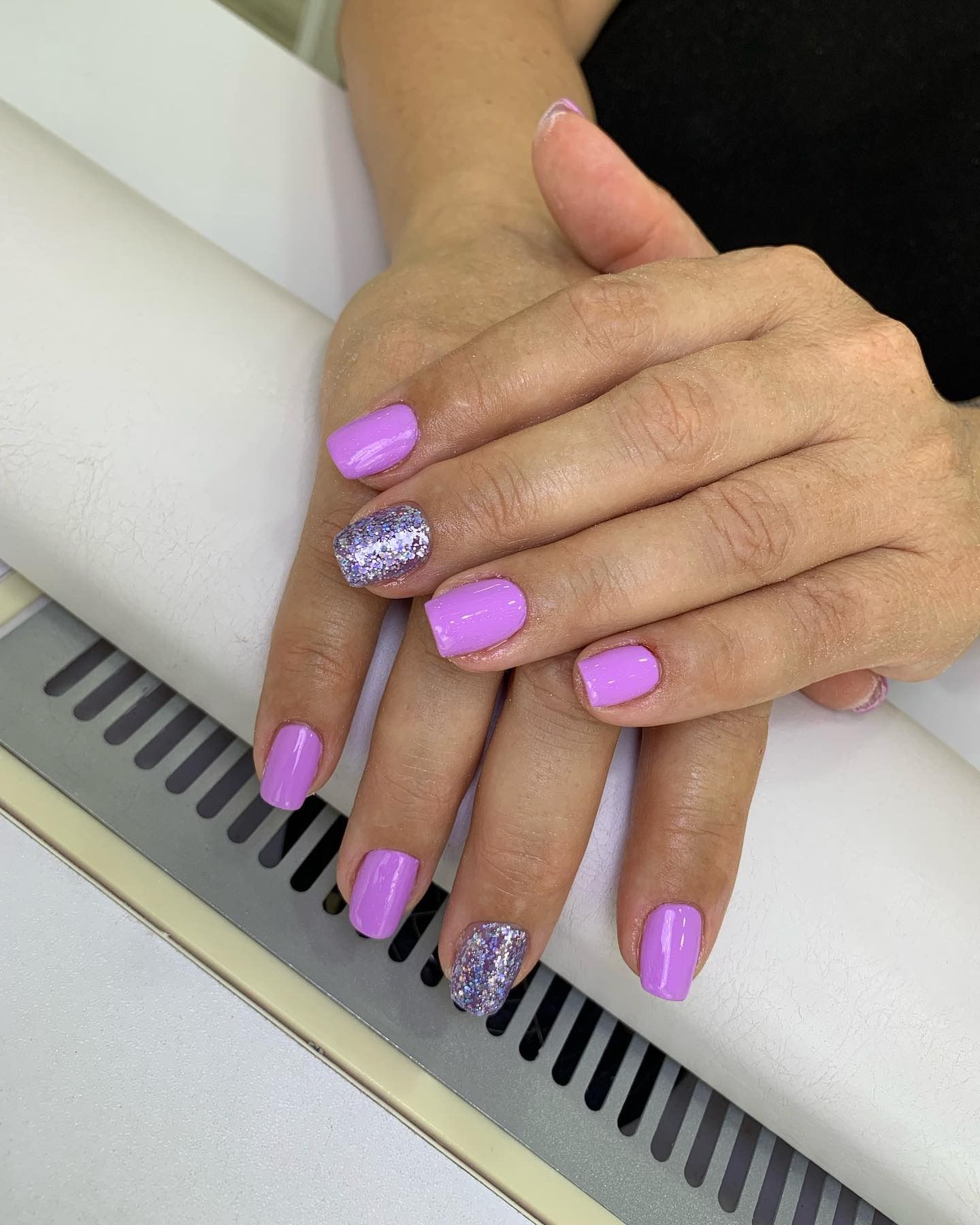Mani con unghie corte color lavanda, due unghie glitterate, appoggiate su una superficie bianca.