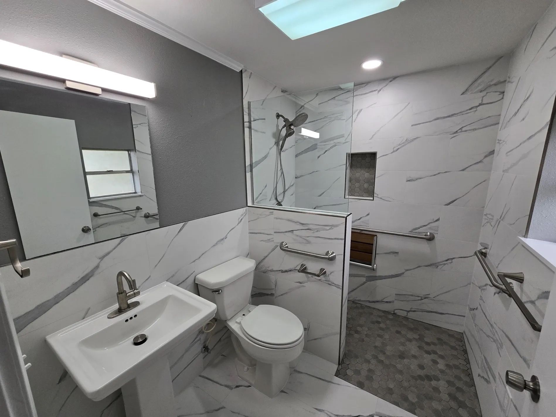 Modern bathroom with marble-look tile, shower, toilet, and sink with grab bars, and a skylight.