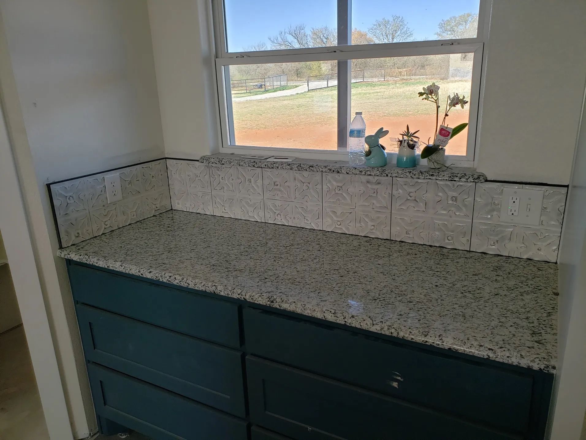 Countertop with cabinets below a window; speckled gray surface with white tile backsplash and teal cabinets.