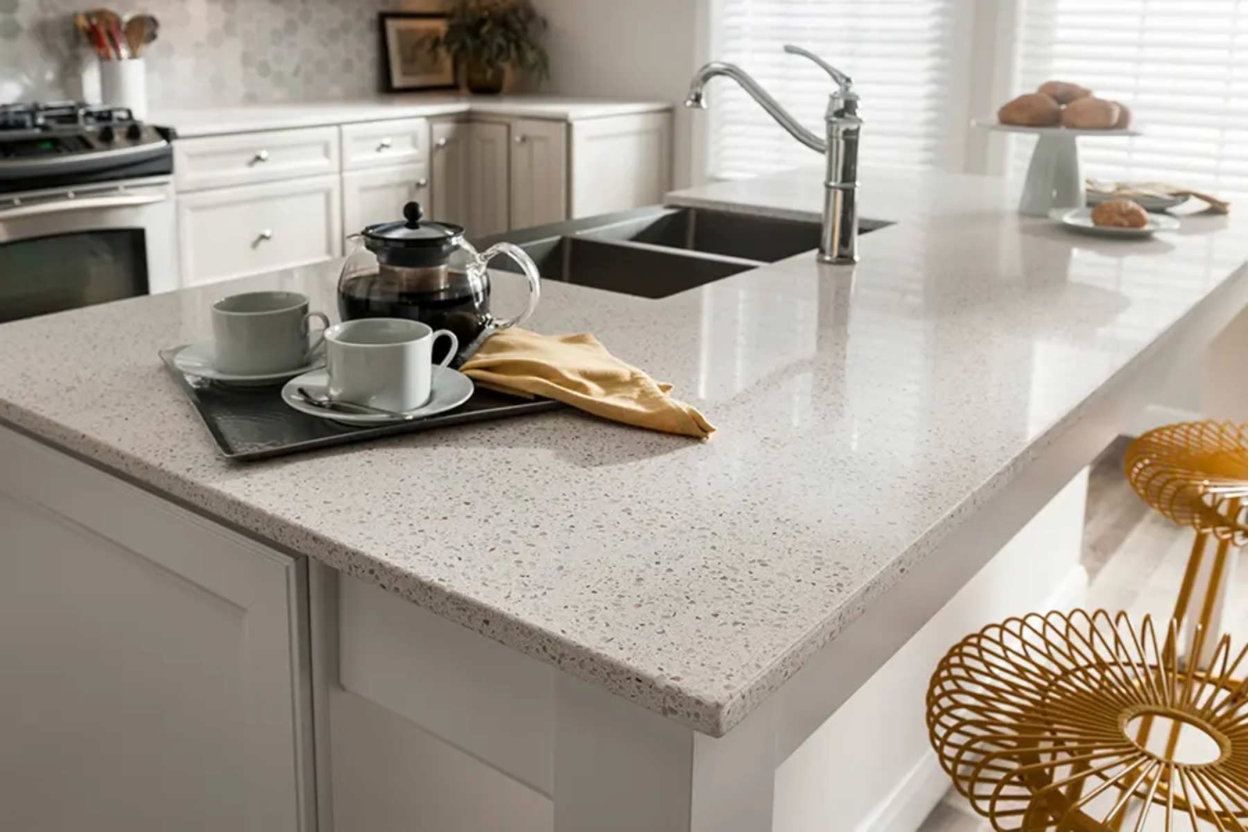 Bright kitchen with a speckled island, sink, coffee cups, teapot, and gold chairs by the counter.