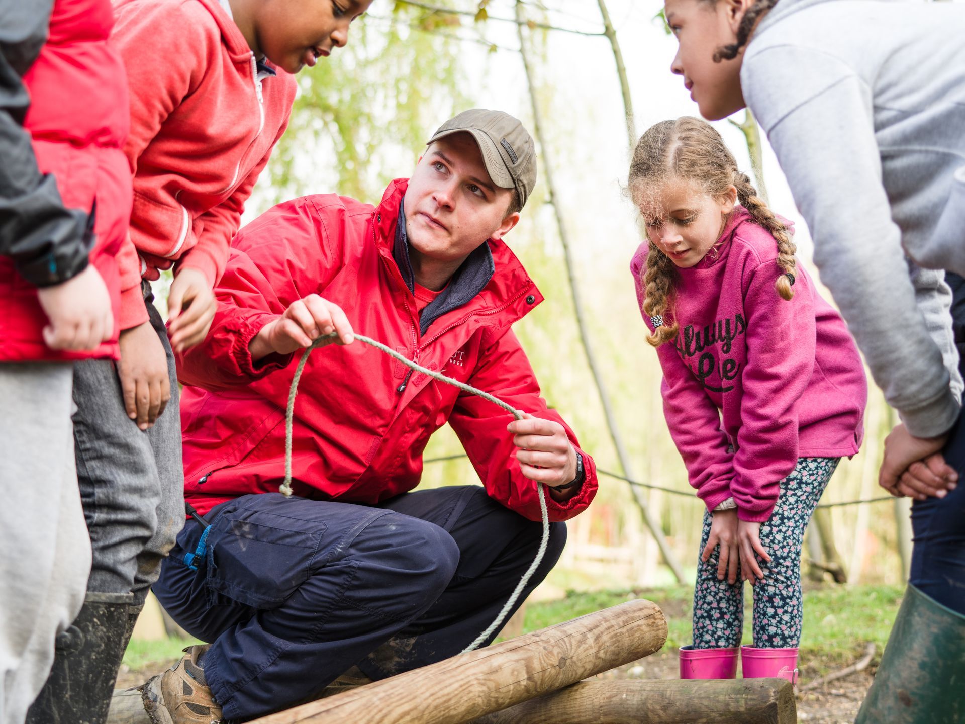 A man in a red jacket is teaching a group of children how to build a fire.