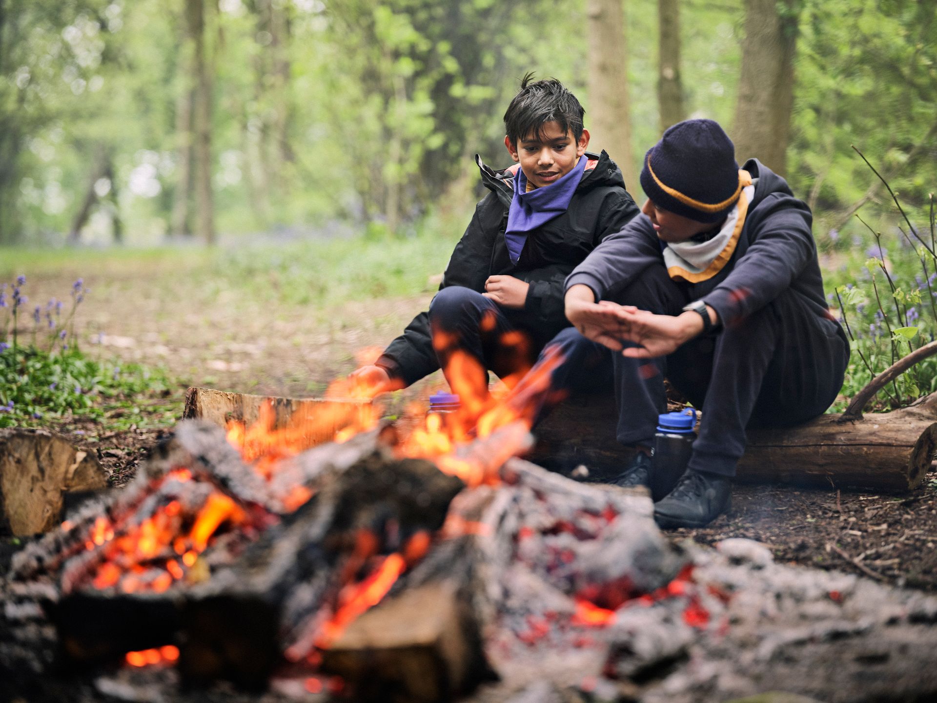 Two young boys are sitting around a campfire in the woods.