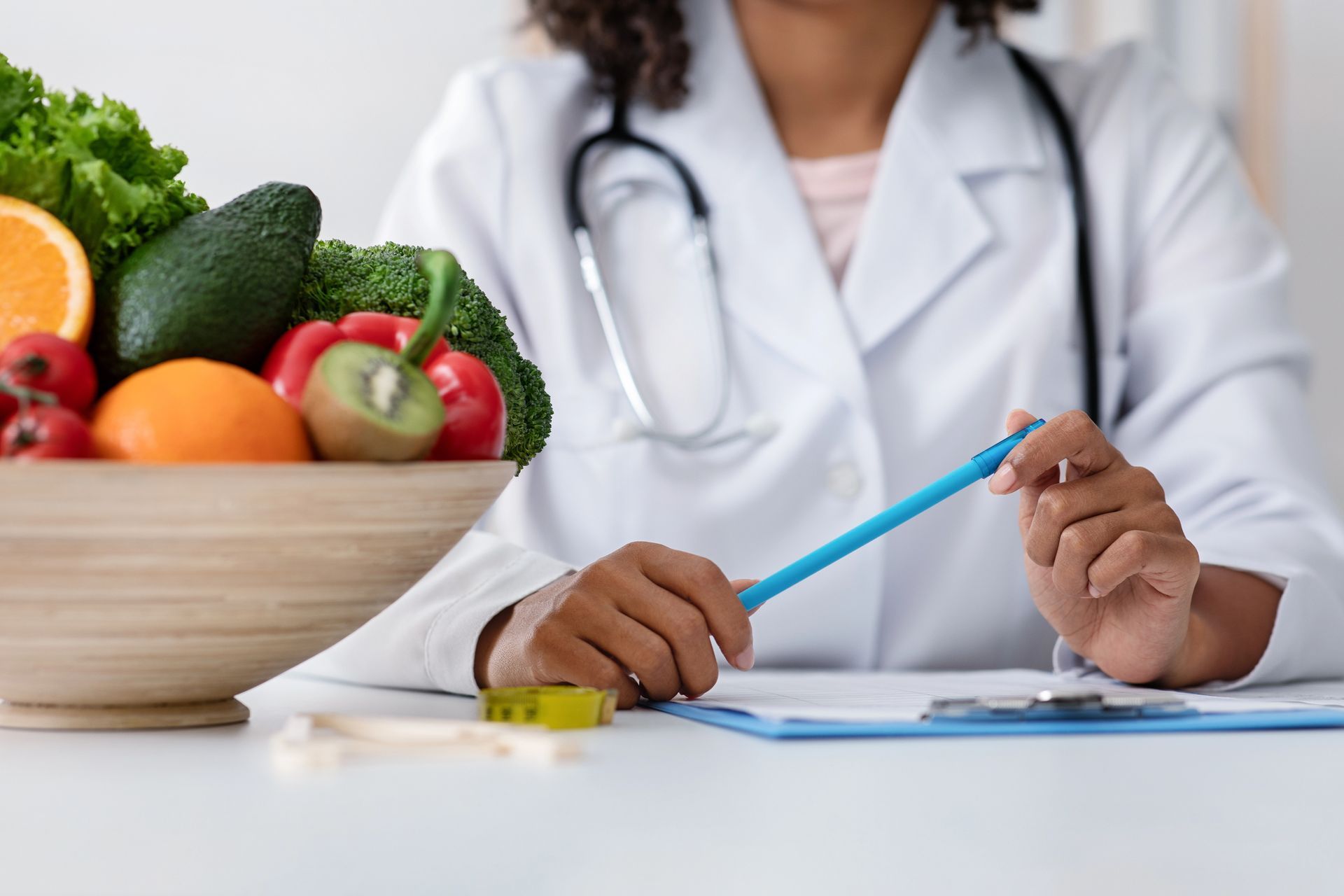 Un profesional sanitario con bata de laboratorio está sentado en un escritorio con un cuenco de fruta y verdura fresca y un portapapeles.