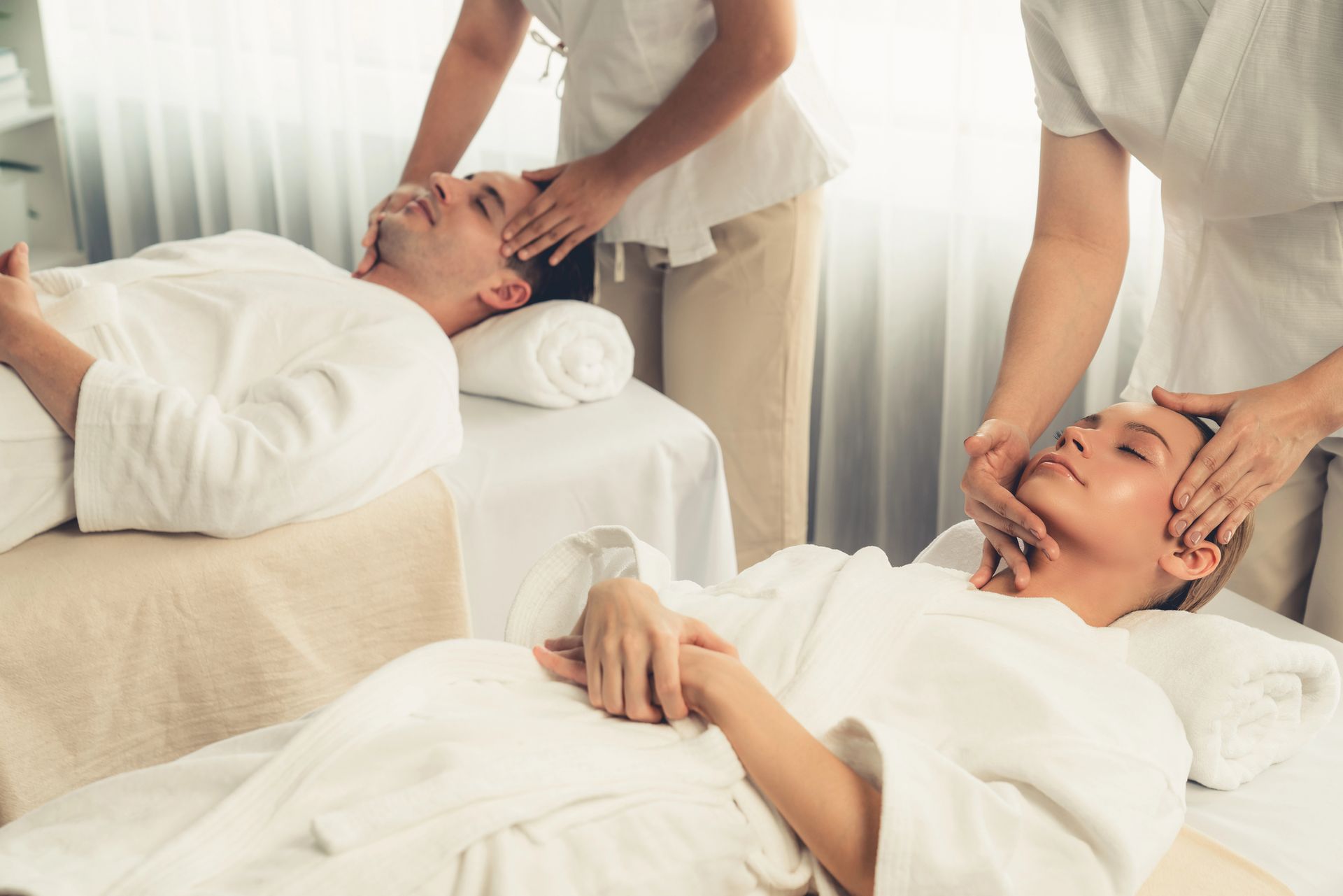 Dos personas con batas blancas yacen tumbadas en camillas de spa mientras reciben masajes faciales del personal en una habitación luminosa y minimalista.