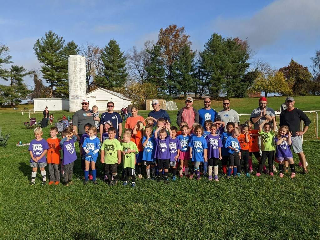 Group of children and adults posing on a grassy field with soccer goals and trees in the background