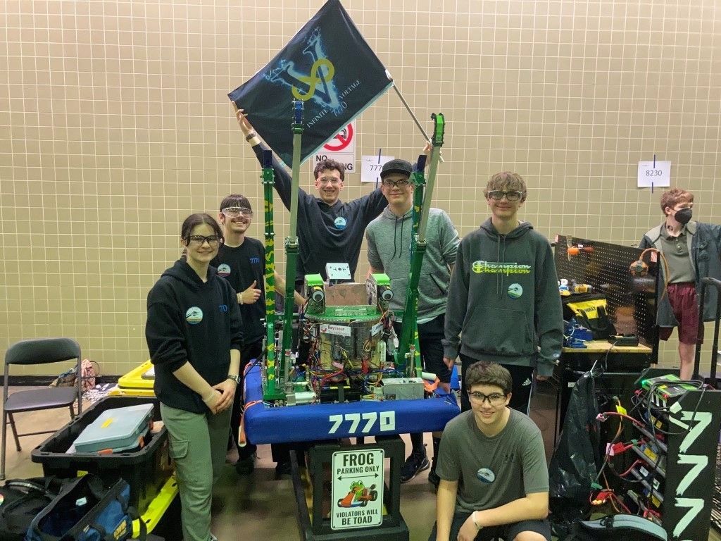 Team of students posing with a green-and-blue robot at a robotics competition, holding a team flag.