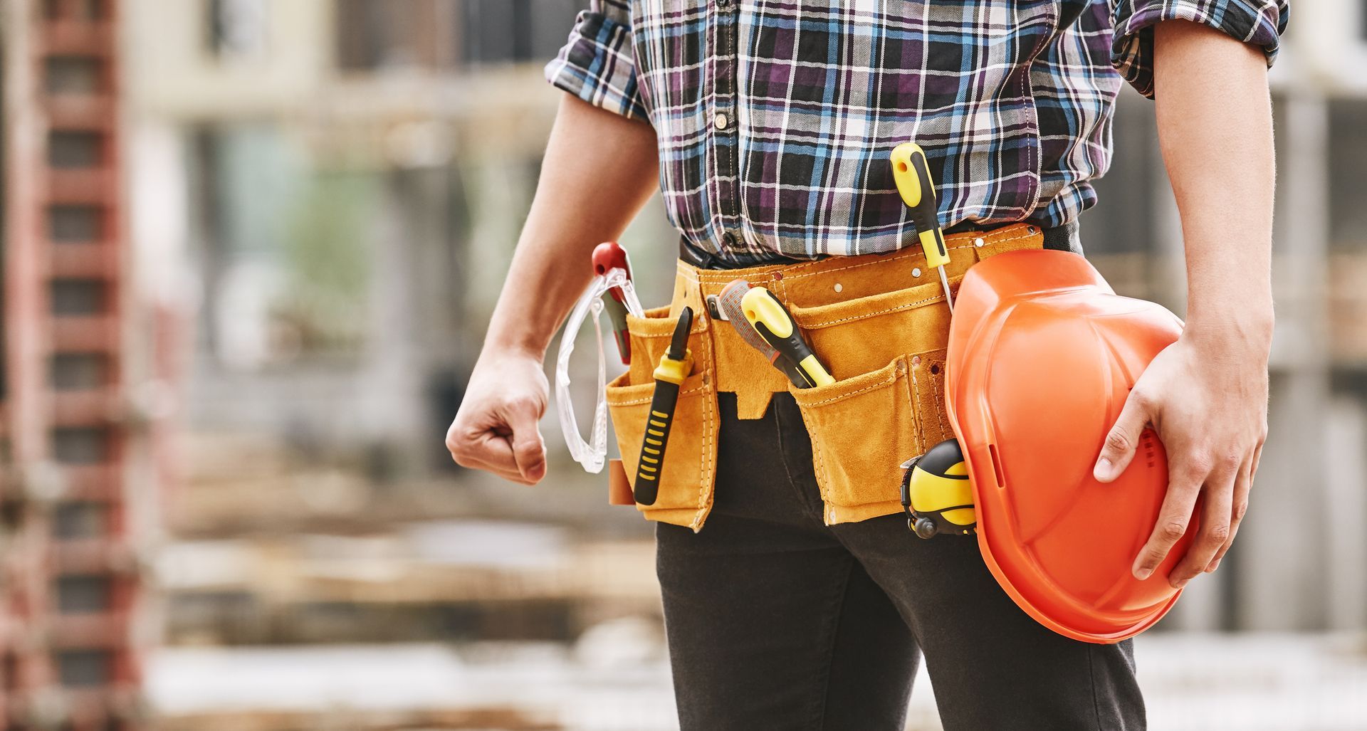 A construction worker is holding an orange hard hat and a tool belt.