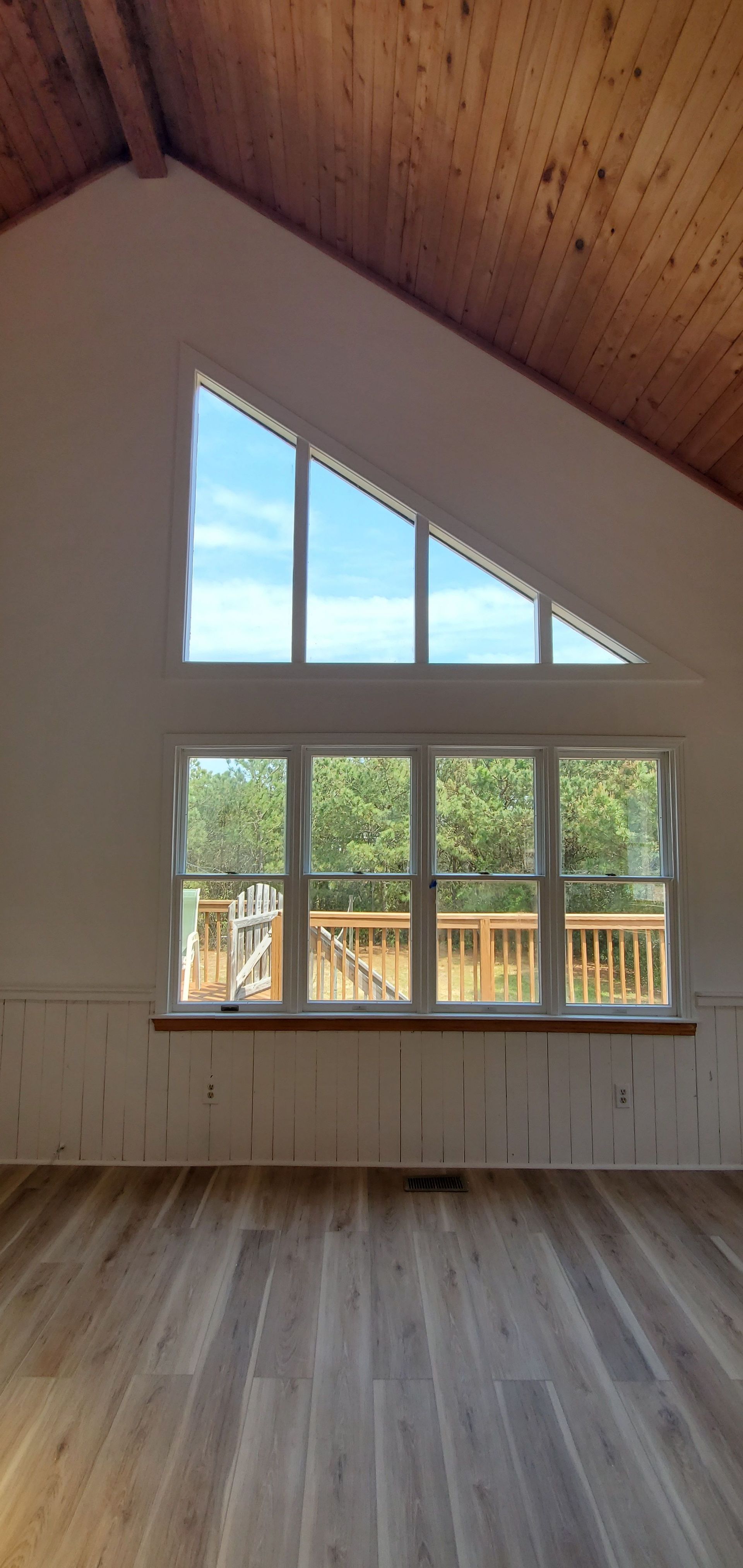Empty loft with wood ceiling and floor, large triangular and rectangular windows overlooking trees.