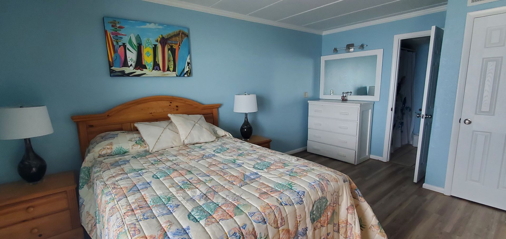 Bright bedroom with a patterned bedspread, wooden dresser, nightstands, and blue walls with an open doorway