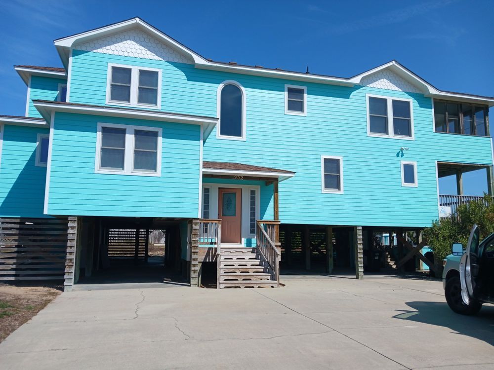 Bright turquoise elevated house with white trim, stairs, and parking area under a blue sky