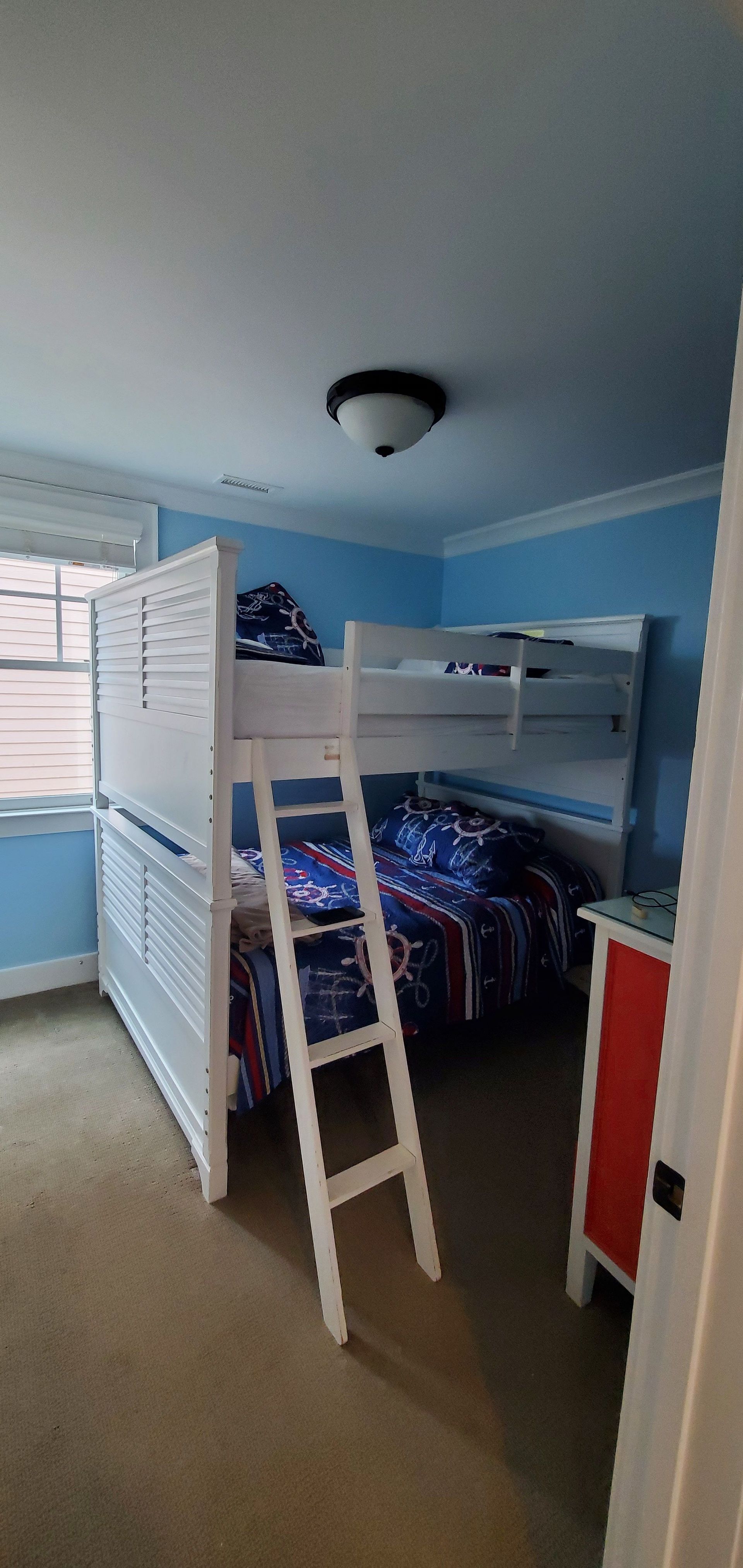 Bright blue bedroom with a white bunk bed, desk, and storage cabinets under a ceiling light.