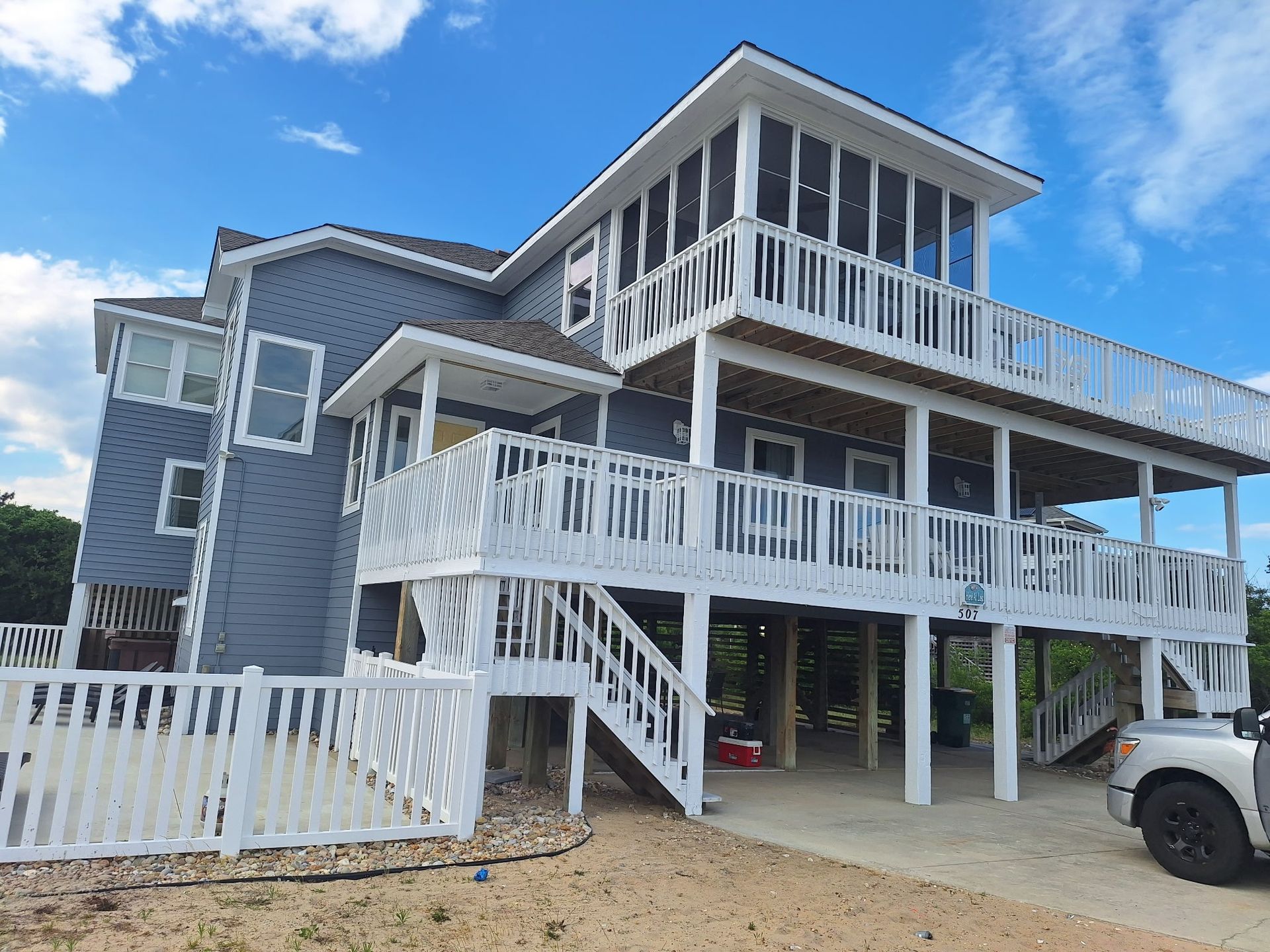 Stilted gray beach house with white railings and stairs under a blue sky, with a white truck parked beside it