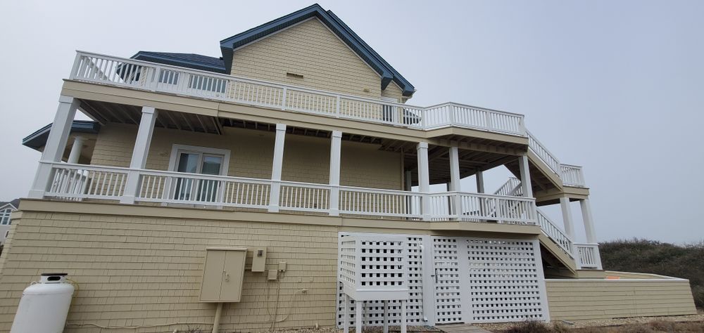 Large beige beach house with wraparound porch and white railings under a cloudy sky.