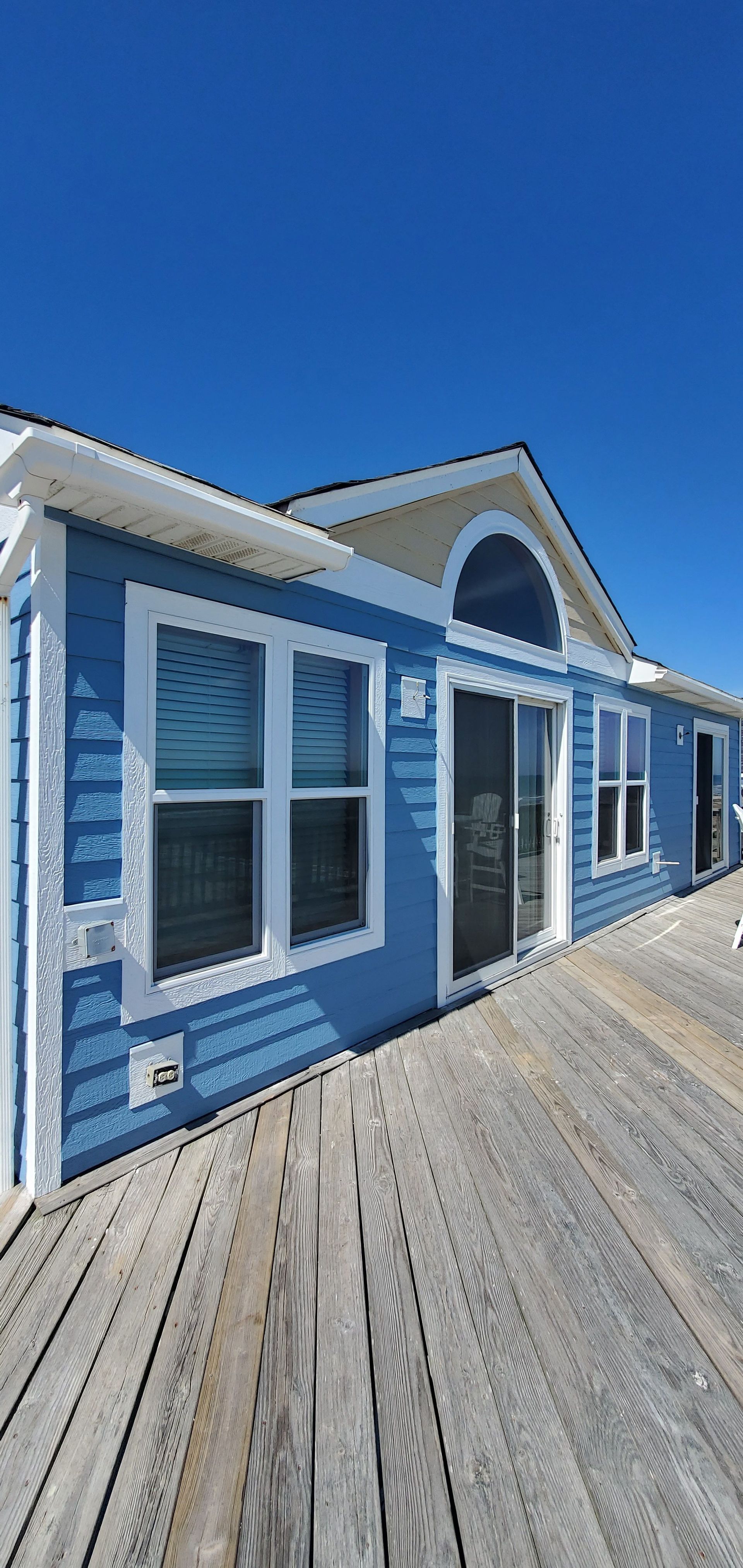 Blue seaside house with white trim and a wooden deck under a bright sky