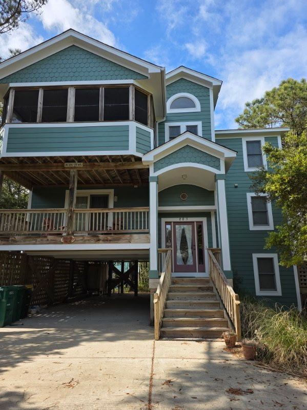 Blue beach house on stilts with covered porch, stairs, and open ground-level parking under a partly cloudy sky