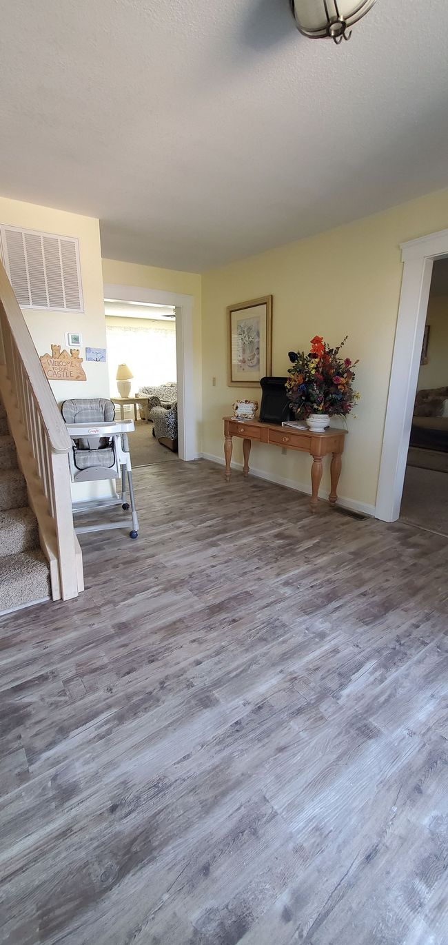 Spacious hallway with wood flooring, white walls, a console table with flowers, and light from a nearby room.