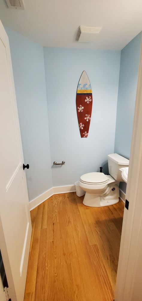 Small bathroom with light blue walls, wood floor, white toilet, and surfboard wall decor.