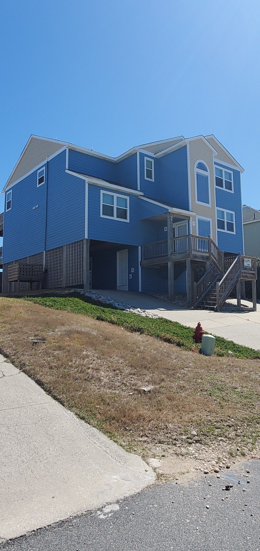 Blue multi-story house on a sloped lot with a gravel driveway and sparse grass under a clear sky