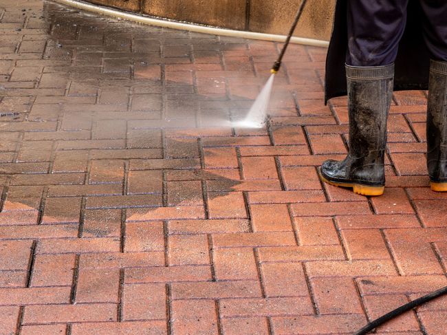 Pressure washing brick pavement, with water spraying across the wet surface and a worker’s boots visible