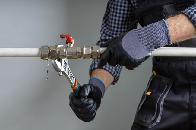 Worker tightening a metal pipe valve with pliers, wearing gloves and a checkered shirt.