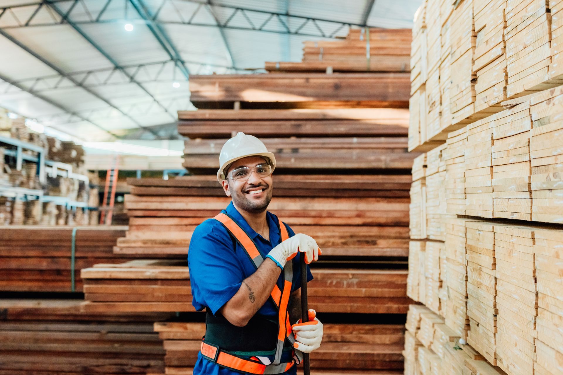 A worker in a lumber warehouse wearing safety gear, holding a tool, with stacks of wood around.