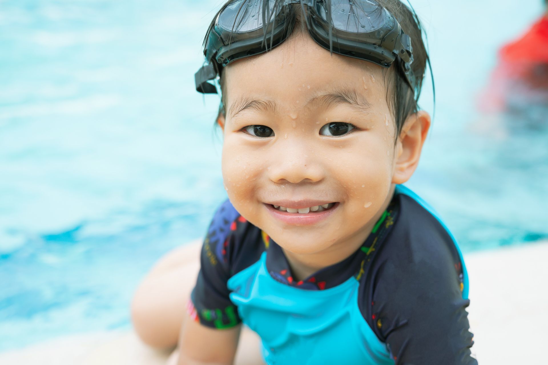 A young boy wearing goggles is sitting on the edge of a swimming pool.