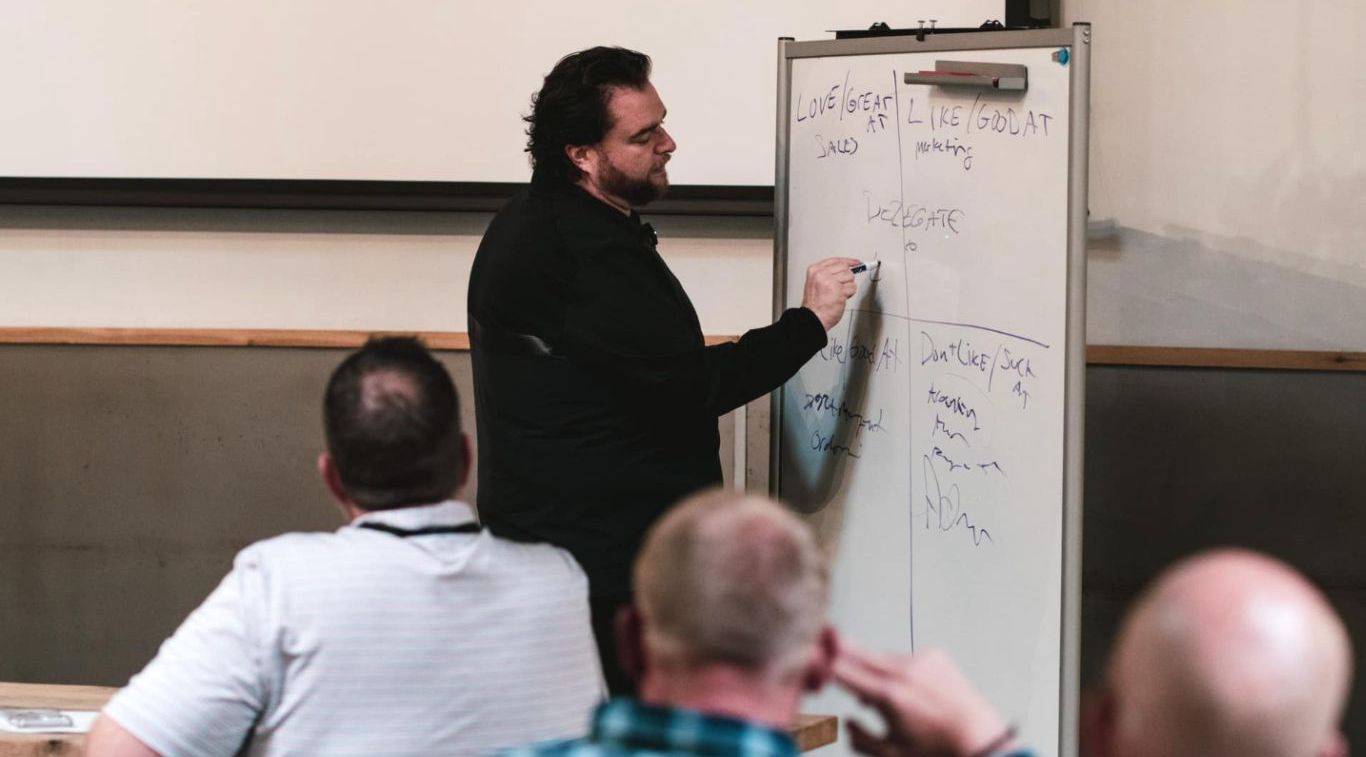 A man is writing on a whiteboard in front of a group of men.