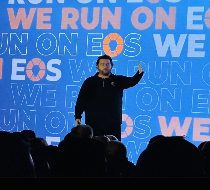 A man stands in front of a blue wall that says we run on eos