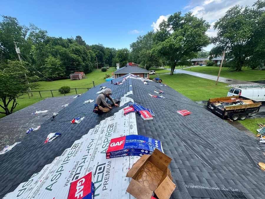Roofer on a dark shingled roof, installing new shingles and underlayment, in a suburban setting.