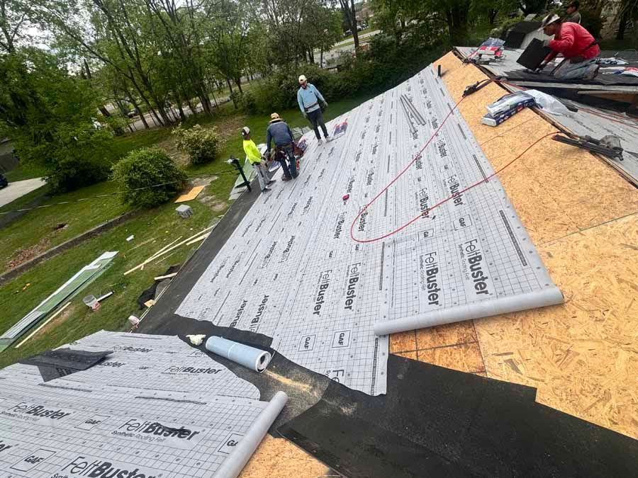 Roofers installing roofing material on a sloped roof, with safety harnesses, green trees and grass in the background.