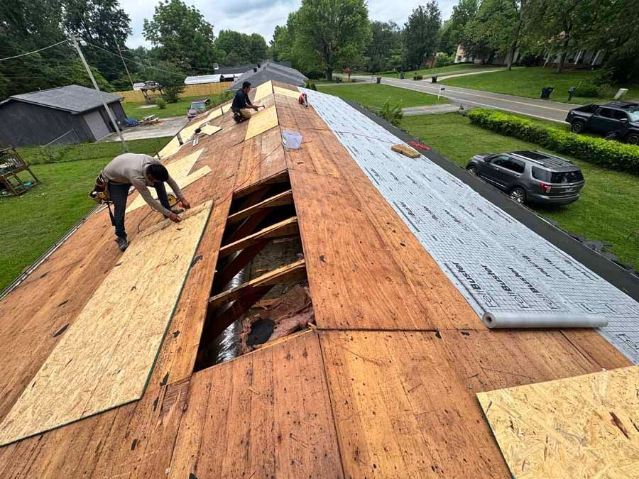 Two roofers replacing a roof, laying plywood. Green grass, gray cars in the background.