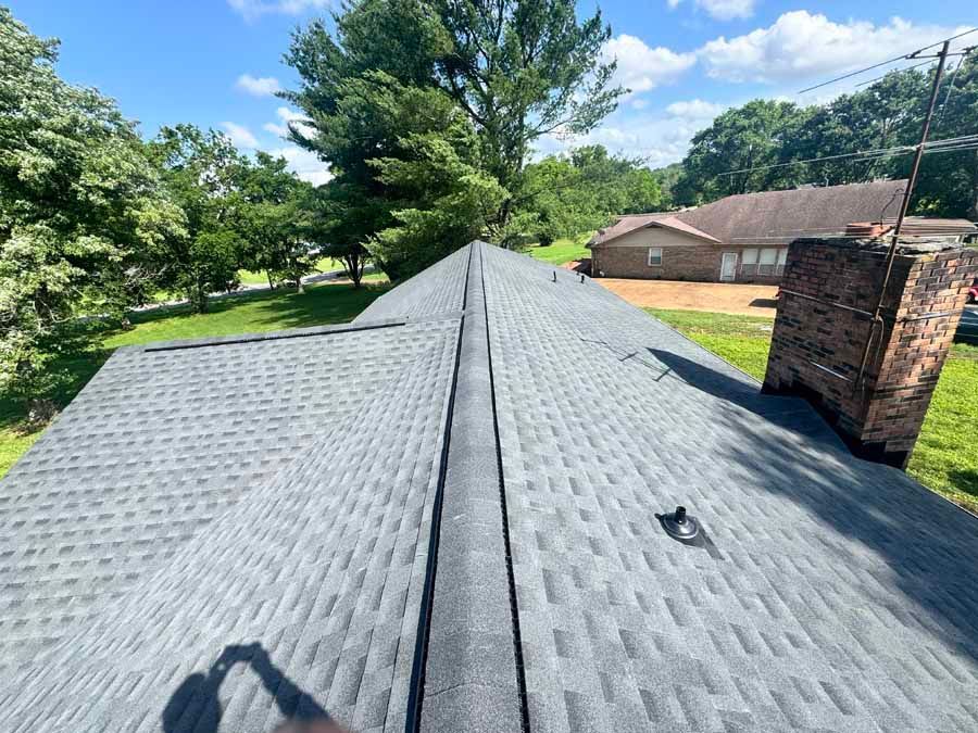 Dark shingle roof with black ridge, brick chimney, and trees under a blue sky.